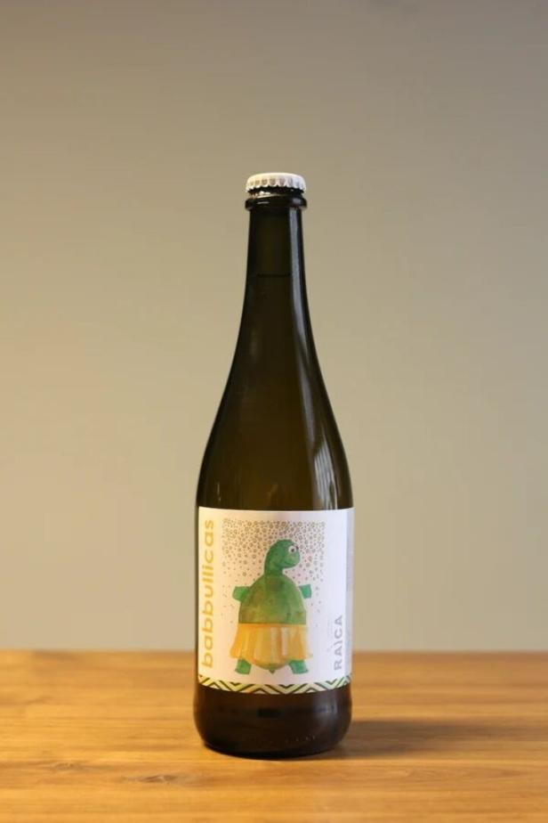 Eine Flasche Babbullicas von Raica mit einem Etikett, das eine grüne Schildkröte in einem gelben Rock zeigt, steht auf einem Holztisch.