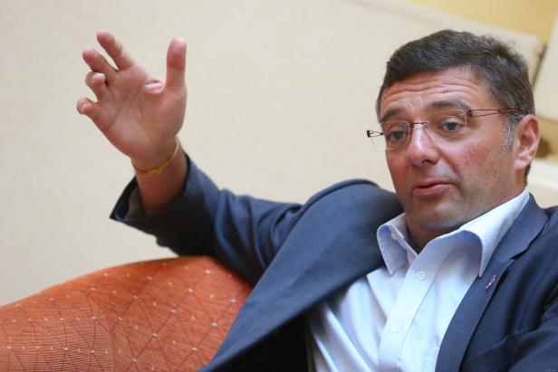 Ein Mann mit Brille und Anzug gestikuliert auf einem orangefarbenen Sessel.