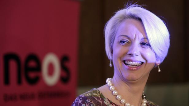 Eine lächelnde Frau mit kurzem, blondem Haar und einer Perlenkette vor einem NEOS-Logo.