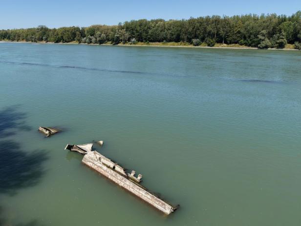 Das Wrack eines gesunkenen Schiffes liegt teilweise unter Wasser in einem breiten Fluss vor einem bewaldeten Ufer.