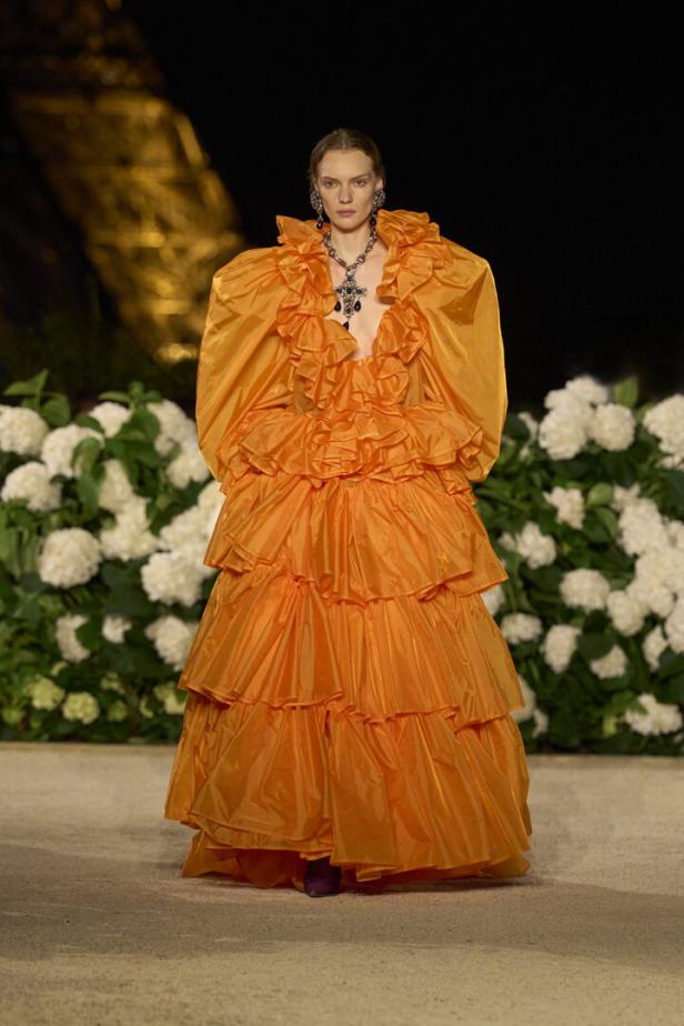Ein Model präsentiert ein voluminöses, orangefarbenes Kleid mit Rüschen vor einer Kulisse aus weißen Hortensien.