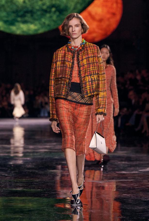 Ein Model präsentiert auf einem Laufsteg ein kariertes Ensemble in Orange- und Brauntönen mit einer weißen Handtasche.