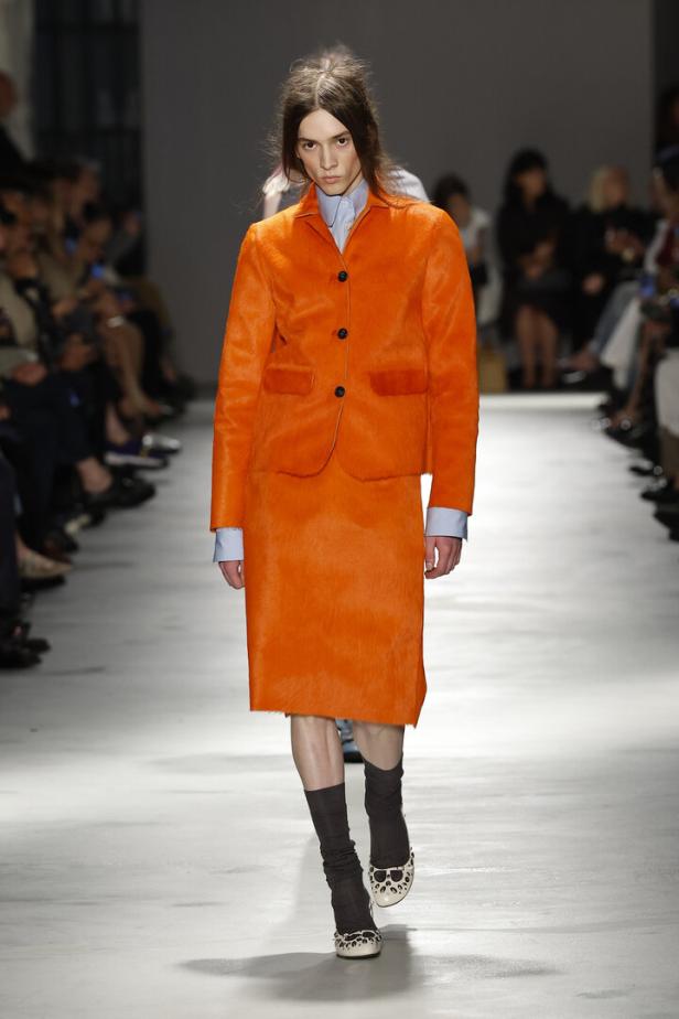 Ein Model präsentiert auf einem Laufsteg ein leuchtend orangefarbenes Kostüm mit hellblauem Hemd, dunklen Socken und gemusterten Schuhen.