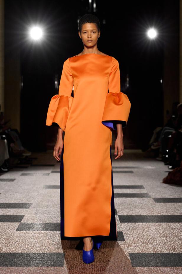 Ein Model präsentiert auf einem Laufsteg ein langes, orangefarbenes Kleid mit weiten Ärmeln und violetten Akzenten sowie passenden blauen Schuhen.