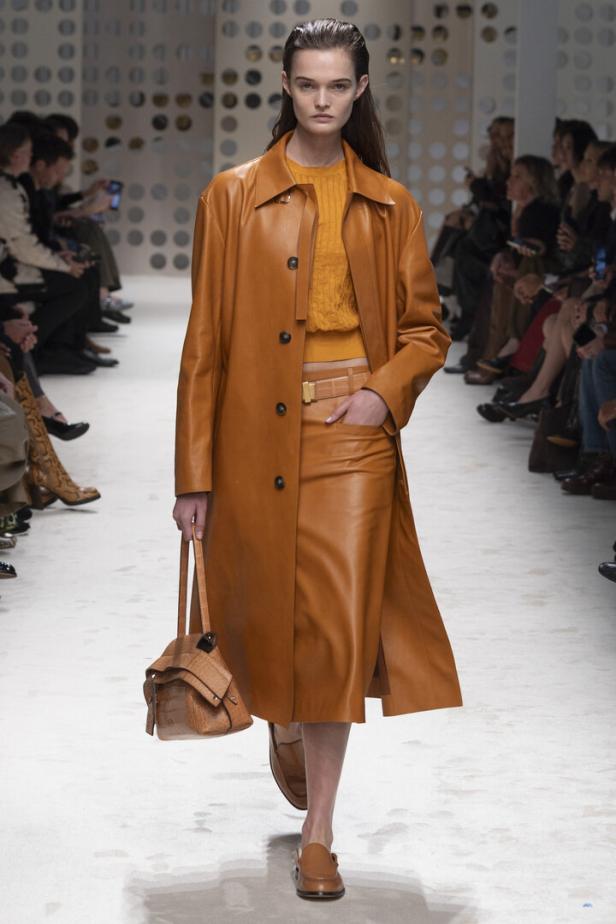 Ein Model präsentiert auf einem Laufsteg einen orangefarbenen Ledermantel mit passendem Rock, gelbem Oberteil und einer Handtasche.