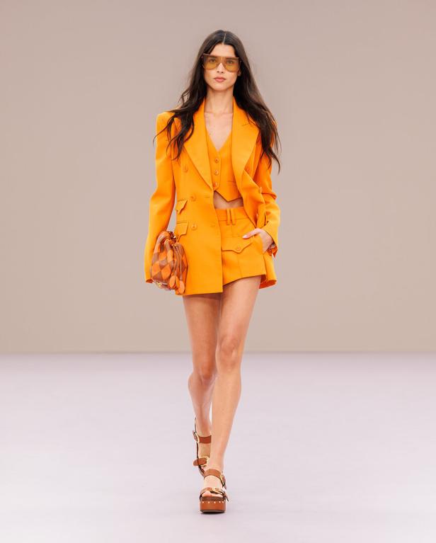 Ein Model schreitet in einem leuchtend orangefarbenen Ensemble aus Blazer, Weste und Shorts mit einer gemusterten Tasche über den Laufsteg.