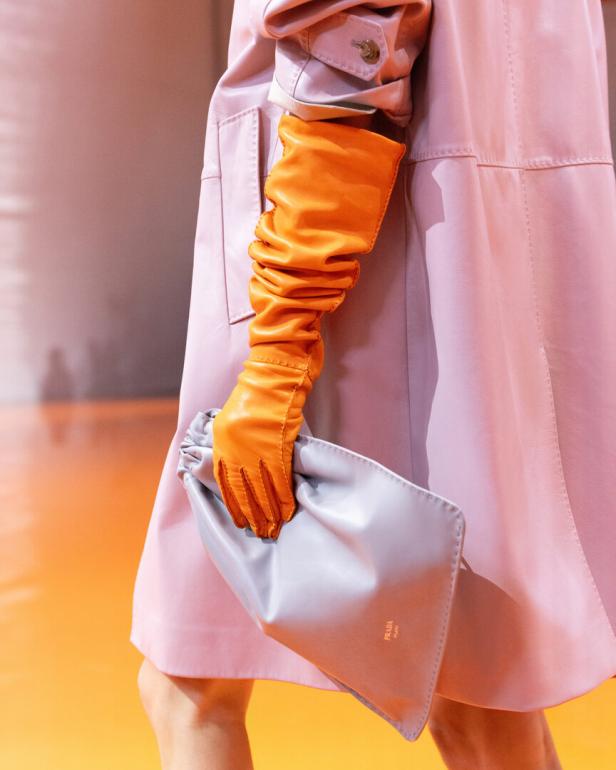 Eine Person in einem hellrosa Ledermantel trägt einen langen orangefarbenen Handschuh und hält eine hellgraue Prada-Clutch in der Hand.