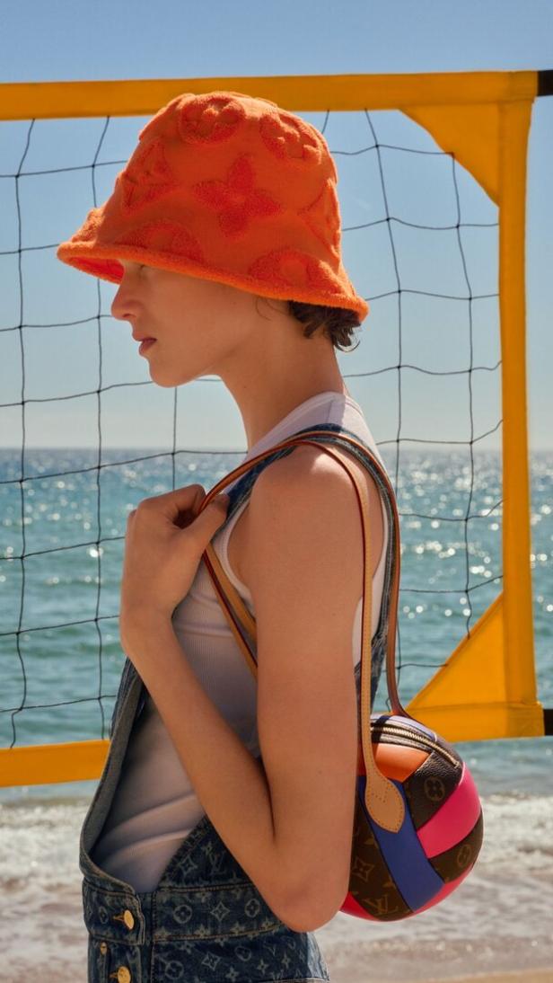 Eine Person mit orangefarbenem Louis Vuitton-Frottee-Fischerhut trägt eine bunte, ballförmige Tasche vor einem Volleyballnetz am Strand.