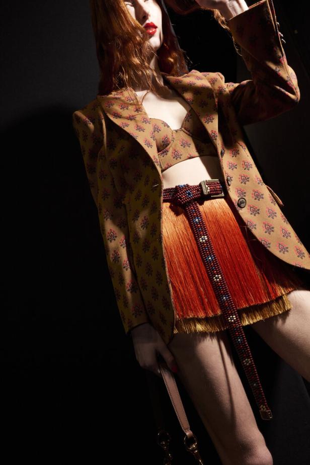 Eine rothaarige Frau trägt einen gemusterten Blazer über einem Bustier und einen orangefarbenen Fransenrock mit einem verzierten Gürtel.