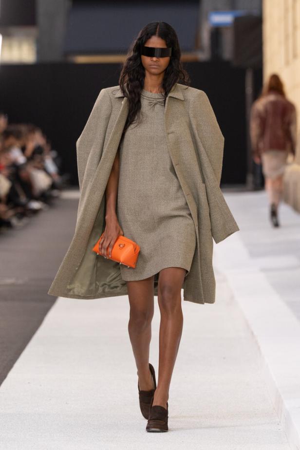 Model auf dem Laufsteg in beigem Mantel und Kleid, mit orangener Clutch und schwarzer Augenbedeckung.