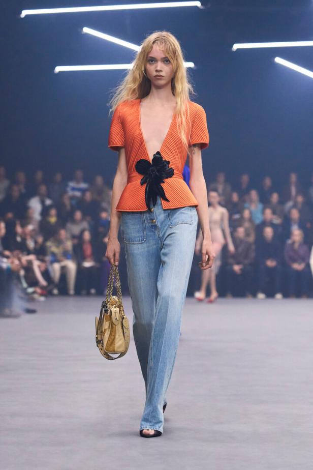 Model auf dem Laufsteg in orangefarbener Wickelbluse, Jeans und mit Handtasche.