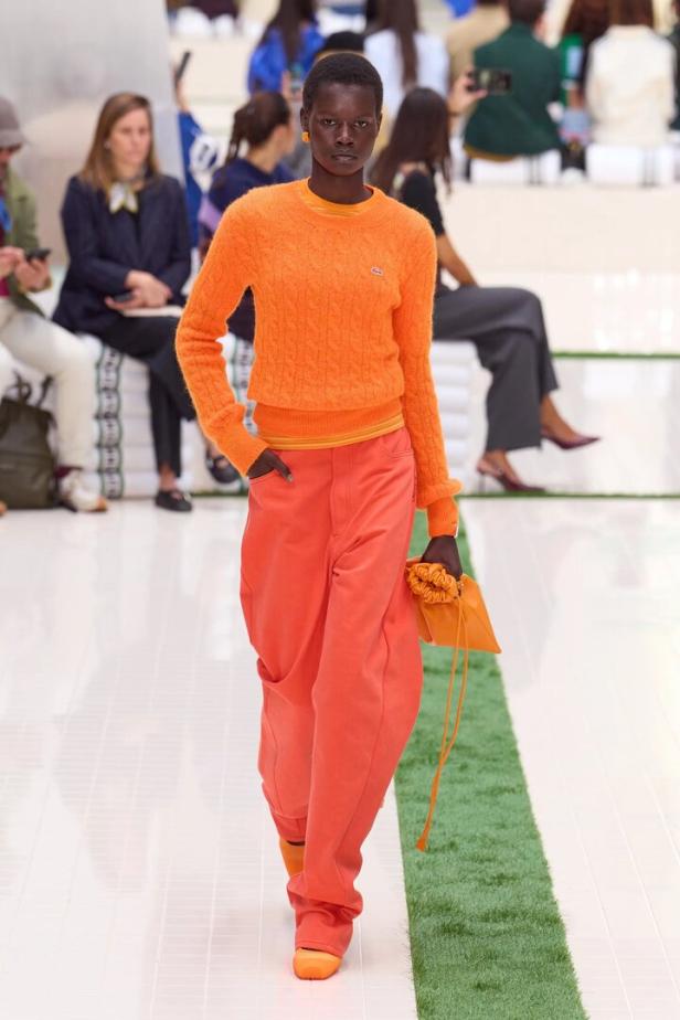 Model auf dem Laufsteg in orangefarbenem Pullover und korallenfarbenen Hosen, mit kleiner Tasche in der Hand.