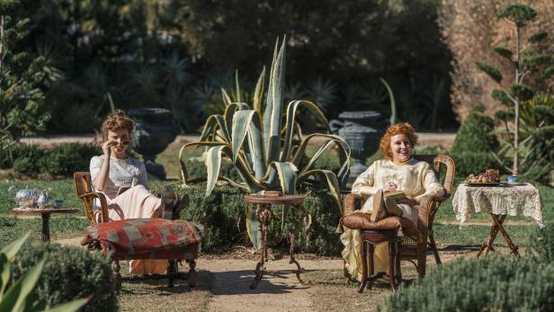 Zwei Frauen sitzen entspannt in einem Garten zwischen großen Agaven und kleinen Tischen.