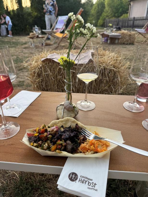 Ein Gericht mit Fleisch und Gemüse auf einer Serviette vom Gasthaus Herbst in Hanfthal, umgeben von Weingläsern und Strohballen im Freien.