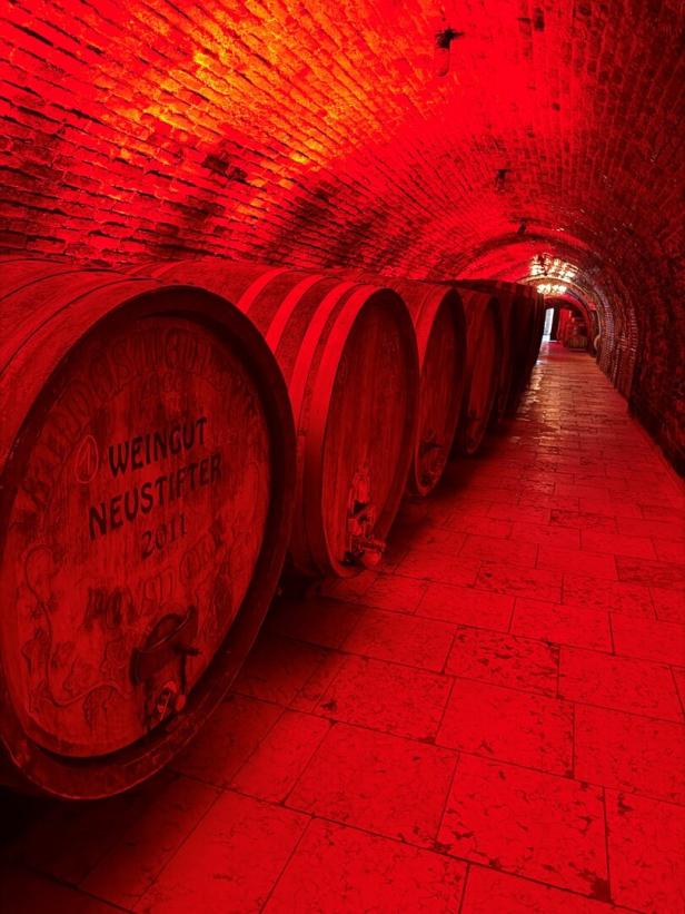 Reihen von Weinfässern des Weinguts Neustifter in einem in rotes Licht getauchten Backsteinkeller.
