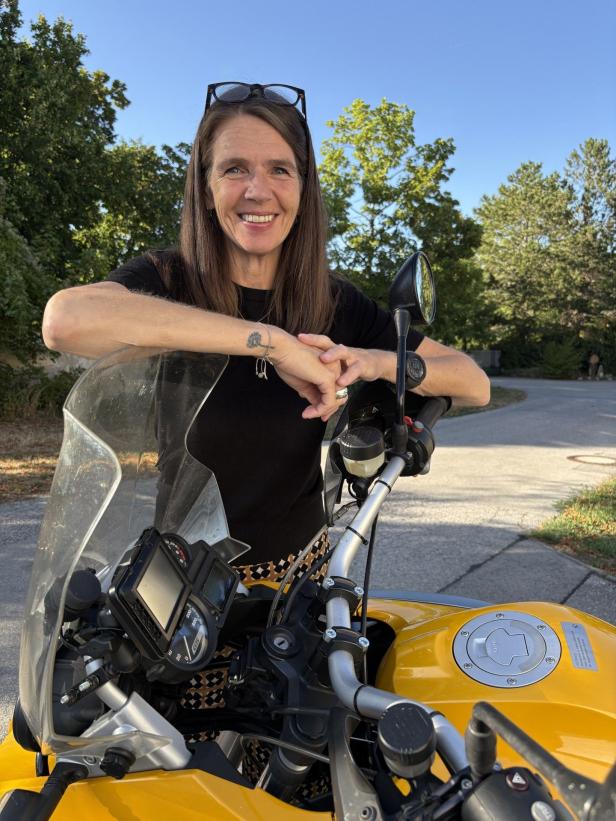 Eine lächelnde Frau lehnt auf einem gelben Motorrad mit Schutzscheibe und steht vor Bäumen unter blauem Himmel.