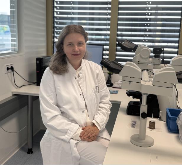 Eine Frau im weißen Laborkittel sitzt im Labor neben einem Mikroskop.
