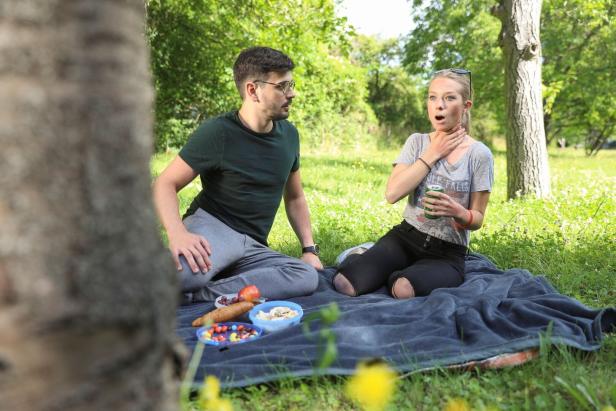 Ein Paar sitzt auf einer Decke im Park; die Frau hält sich erschrocken den Hals, während der Mann sie besorgt ansieht.