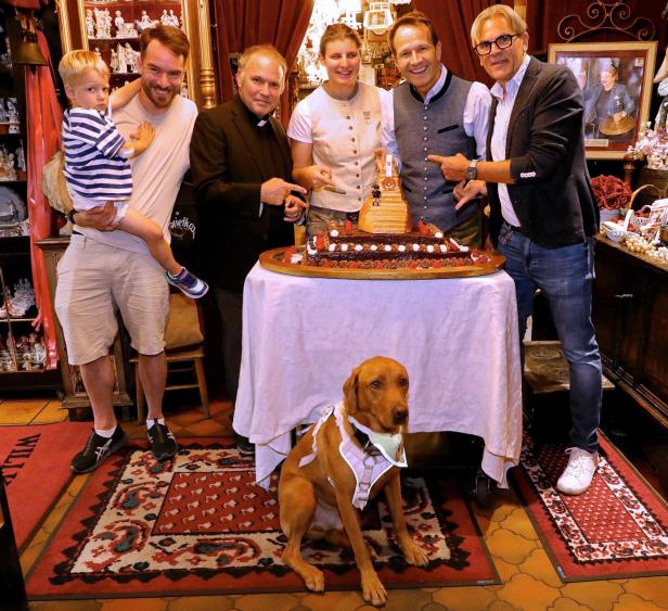 Menschen stehen um einen Tisch mit einer Torte herum, davor sitzt ein Hund