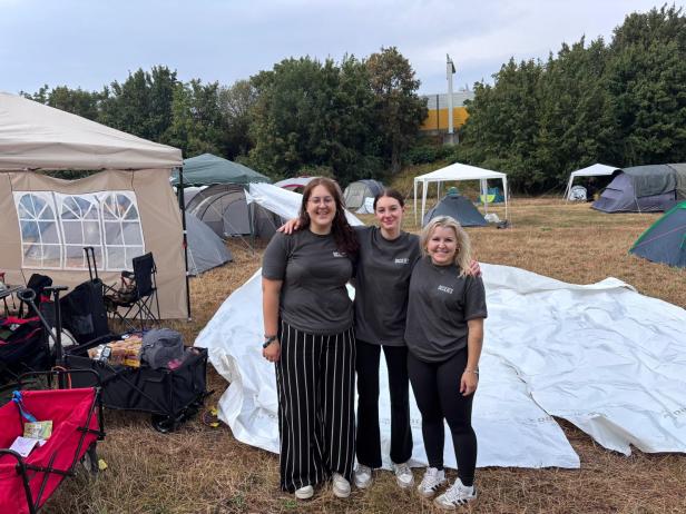 Drei Frauen posieren auf dem Frequency-Campingplatz vor Zelten, Pavillons und Campingausrüstung.