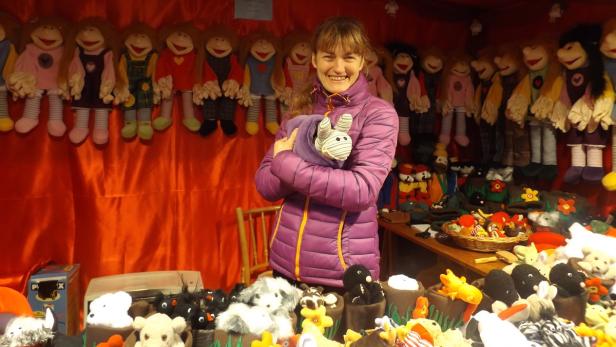 Eine lächelnde Frau steht an einem Stand mit handgemachten Puppen und Stofftieren.