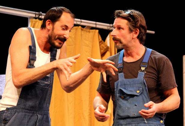 Zwei Männer mit Schnurrbärten und blauen Overalls gestikulieren auf einer Bühne.