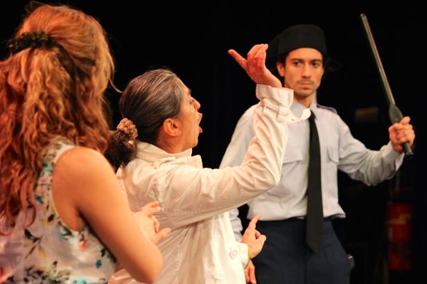 Eine Frau gestikuliert auf der Bühne, während ein Polizist mit Schlagstock zusieht.