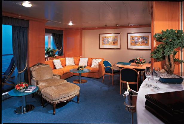 Ein Wohnzimmer mit orangefarbenem Sofa, Sessel und Esstisch auf einem Kreuzfahrtschiff.