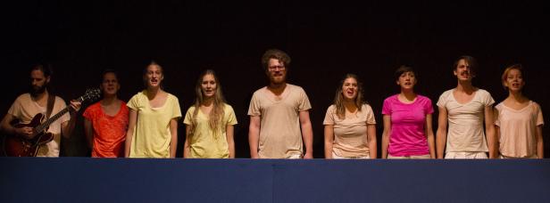 Eine Gruppe von acht Personen steht auf einer Bühne und singt, begleitet von einer Gitarre.