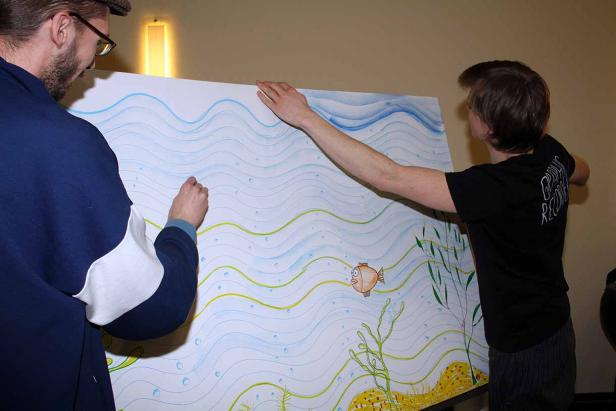 Zwei Personen betrachten eine große Zeichnung mit einem Fisch und Wasserwellen.