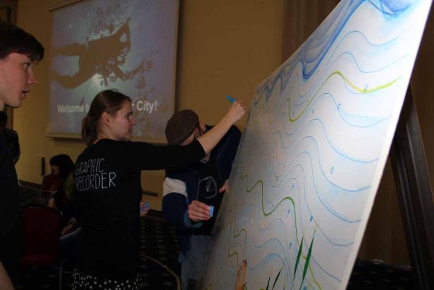 Zwei Personen zeichnen gemeinsam an einem großen, hochformatigen Bild mit Unterwassermotiven.