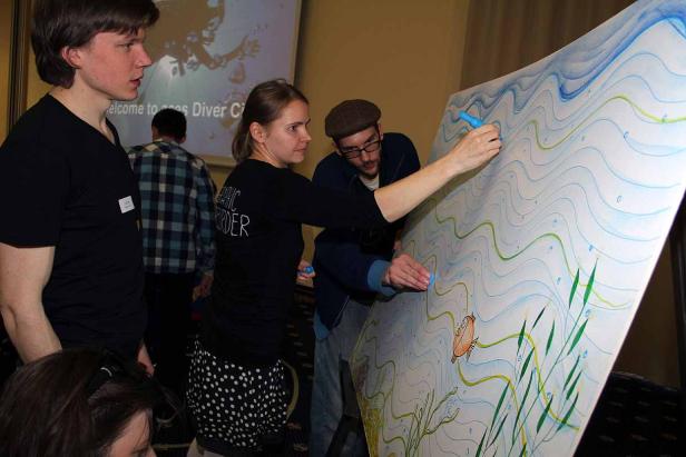 Eine Frau und ein Mann zeichnen gemeinsam ein Unterwassermotiv auf eine große Leinwand.