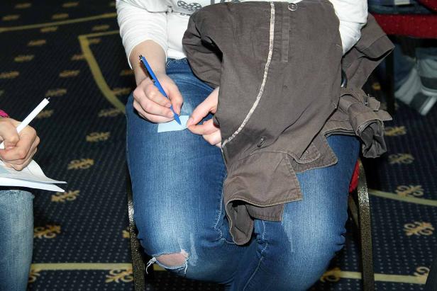 Eine Person sitzt und schreibt mit einem blauen Stift auf einen Zettel, der auf dem Knie liegt.