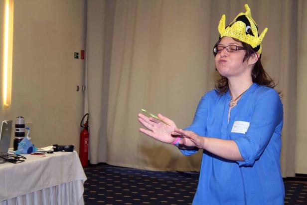 Eine Frau mit Brille und Fischhut jongliert mit einem grünen Gegenstand.