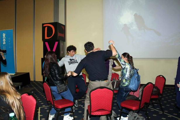 Eine Gruppe junger Leute tanzt in einem Stuhlkreis vor einer Leinwand.