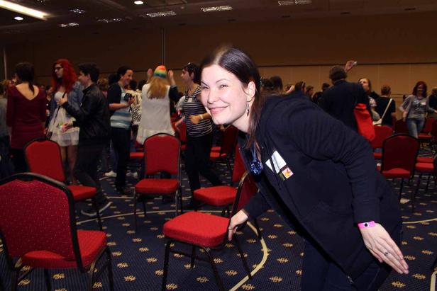 Eine lächelnde Frau steht in einem Raum mit roten Stühlen und vielen Menschen.