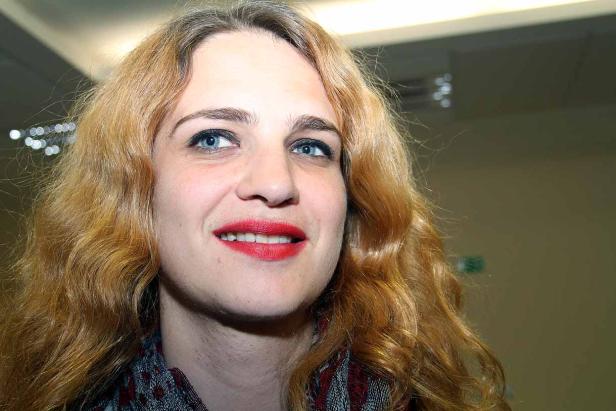 Eine Frau mit blonden, gewellten Haaren und rotem Lippenstift lächelt.