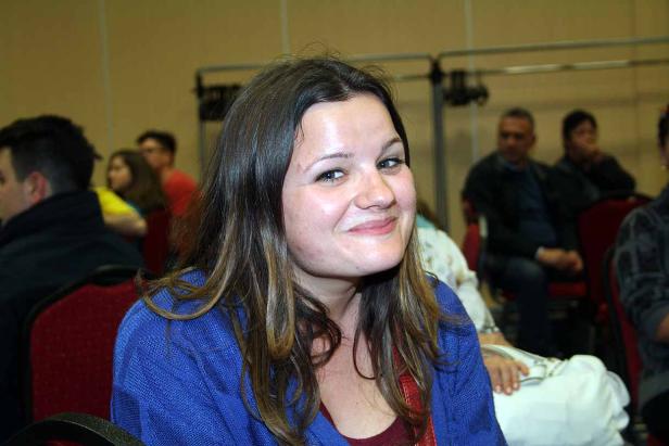 Eine lächelnde Frau mit langen, braunen Haaren sitzt in einem Raum voller Menschen.