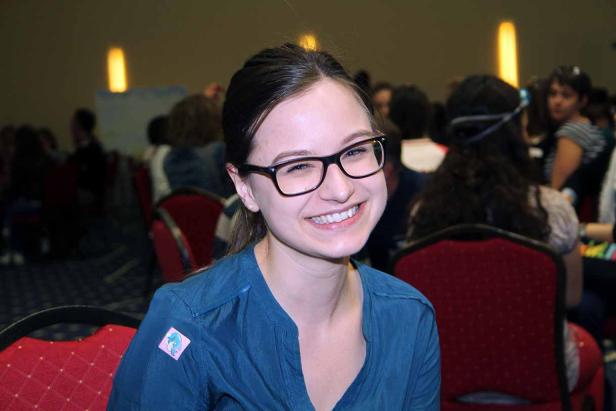 Eine lächelnde junge Frau mit Brille sitzt in einem Raum voller Menschen.
