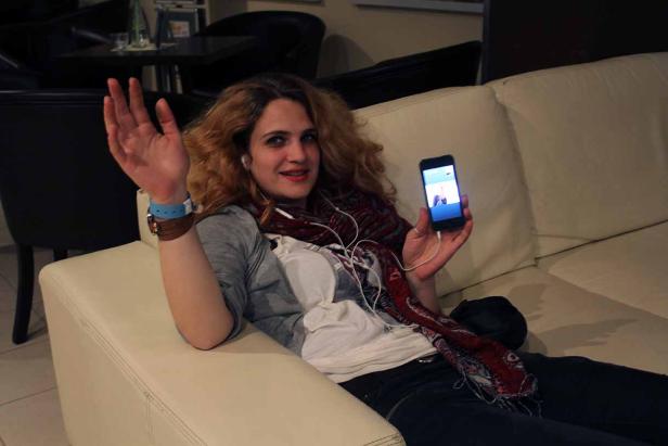 Eine Frau liegt auf einem Sofa, hält ein Smartphone in der Hand und winkt.