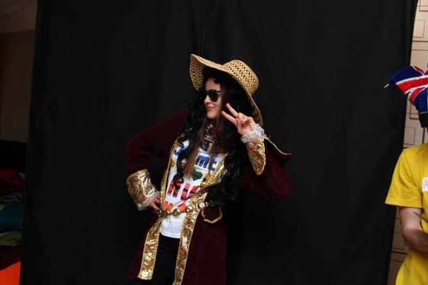 Eine Person mit Sonnenbrille, Hut und auffälligem Kostüm posiert vor einem schwarzen Hintergrund.