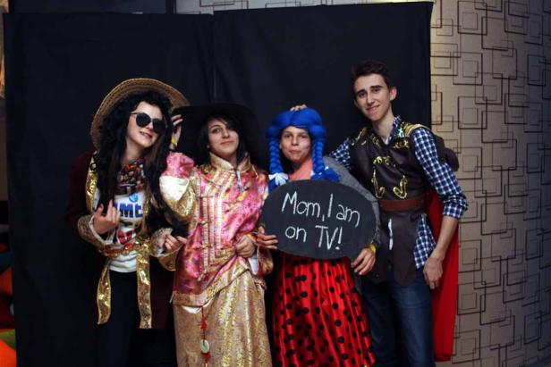 Vier Personen in Kostümen posieren mit einem Schild, auf dem „Mom, I am on TV!“ steht.