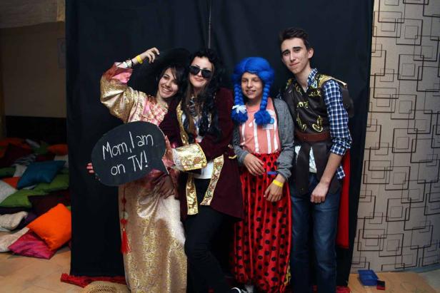 Vier Personen posieren kostümiert vor einer schwarzen Leinwand, eine hält ein Schild mit der Aufschrift „Mom, I am on TV!“.