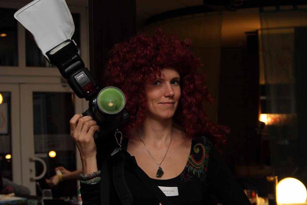 Eine Frau mit roter Lockenperücke hält eine Kamera mit Blitzgerät in der Hand.