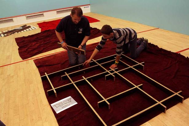 Zwei Personen bauen ein hölzernes Labyrinth auf einem roten Tuch in einer Halle.