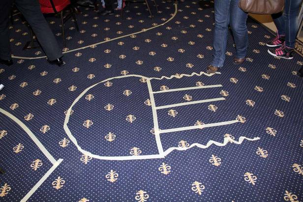 Auf einem Teppich ist mit Klebeband ein stilisierter Kopf gezeichnet.