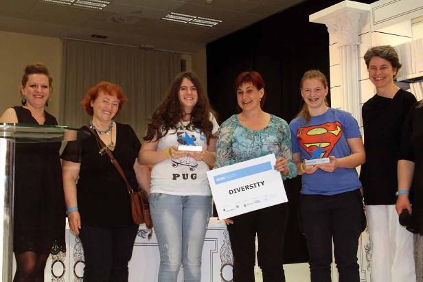 Eine Gruppe von Frauen steht auf einer Bühne und hält Auszeichnungen und ein Schild mit der Aufschrift „Diversity“.