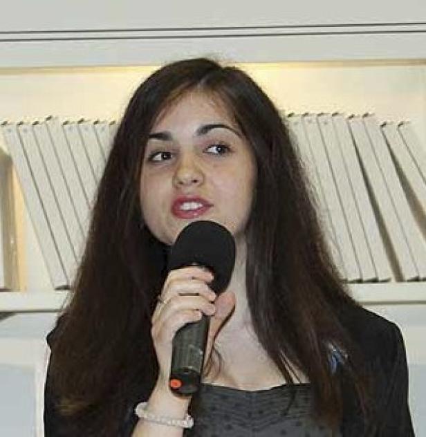 Eine junge Frau mit dunklen Haaren spricht in ein Mikrofon.