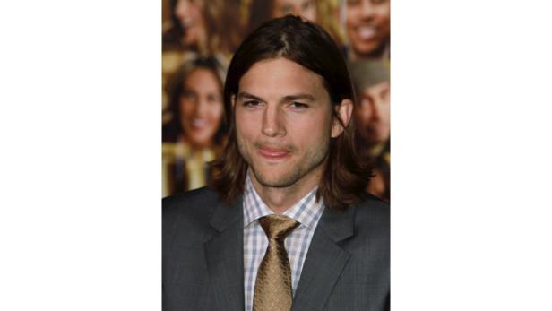 Ashton Kutcher in einem grauen Anzug streckt die Zunge heraus.
