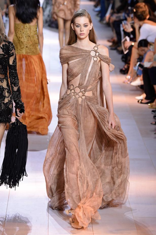 Ein Model präsentiert ein beigefarbenes, transparentes Kleid mit Ringdetails auf dem Laufsteg.